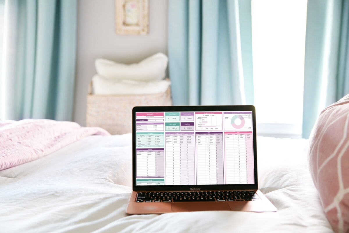 A laptop displaying a colorful spreadsheet—perfect for managing finances or finding budgeting tips—sits on a neatly made bed with white and pink bedding. Light blue curtains and a woven basket with pillows are softly lit in the background.