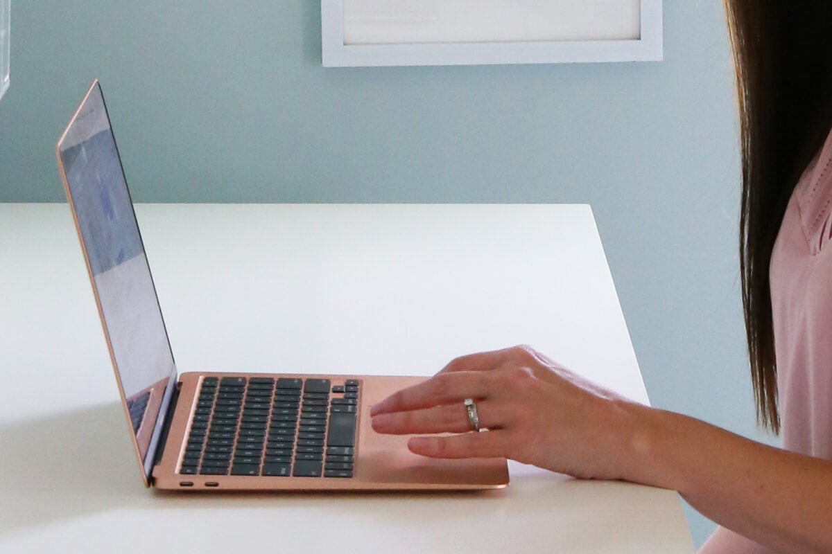 A person with long hair, wearing a light pink shirt and a silver ring, is researching how to budget with irregular income on a slim, gold-colored laptop at a white desk against a pale blue wall. Only their hand and part of their arm are visible.