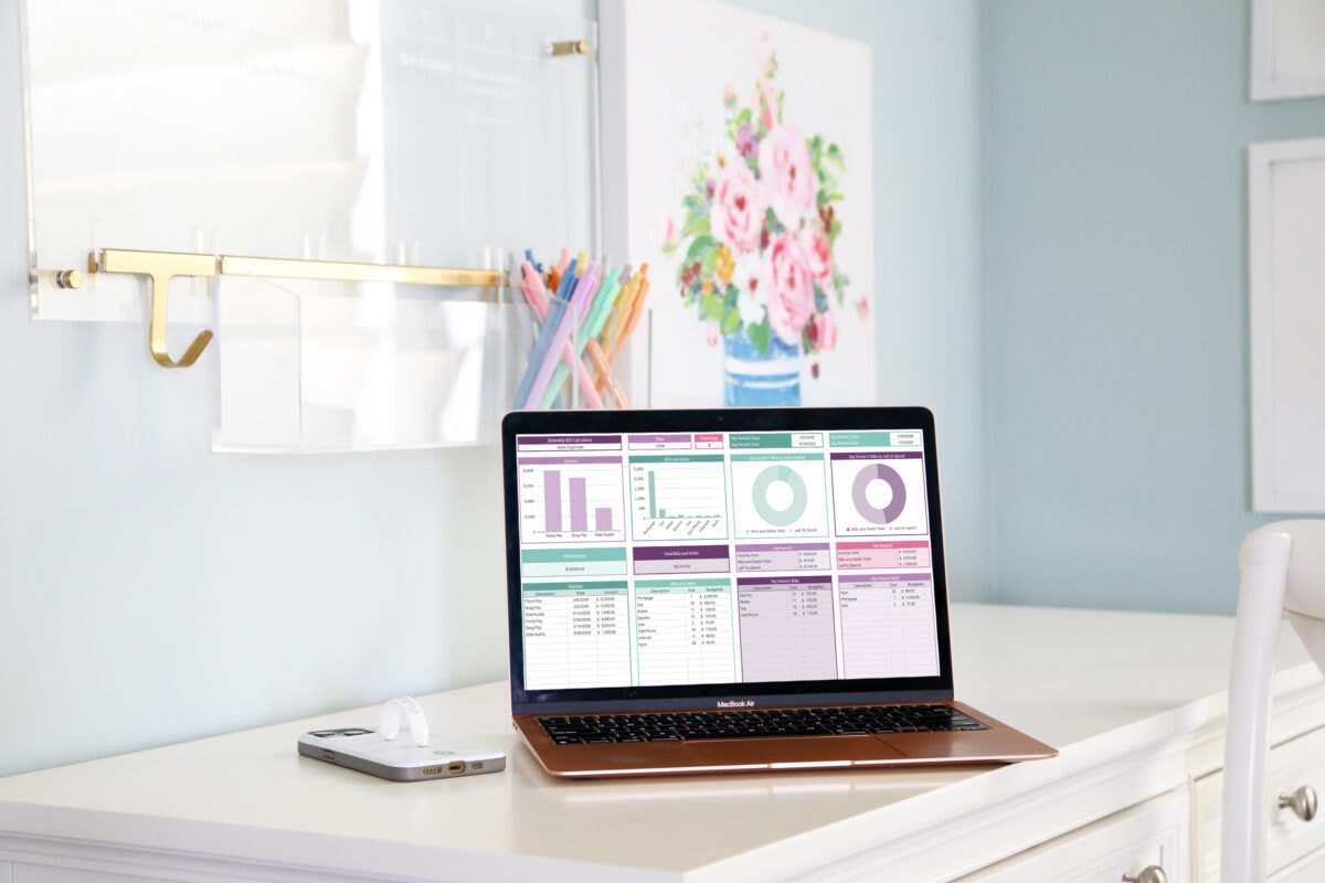 A laptop displaying colorful budgeting charts and graphs sits on a white desk next to a phone and AirPods, with pastel pens in a cup and a floral painting in the background.