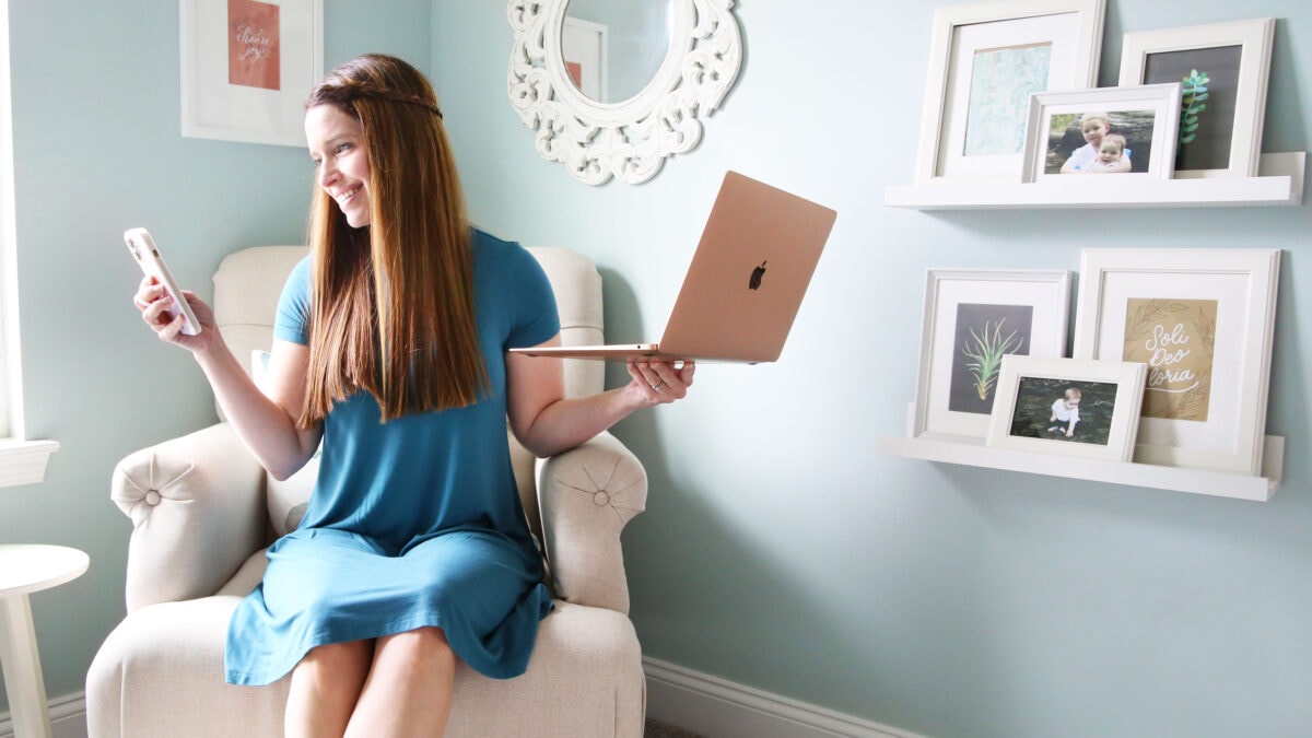 A woman in a blue dress sits on a beige armchair, smiling at her phone while holding a laptop—perhaps exploring budgeting tips. Behind her are white shelves with framed photos and a decorative mirror on a light blue wall.