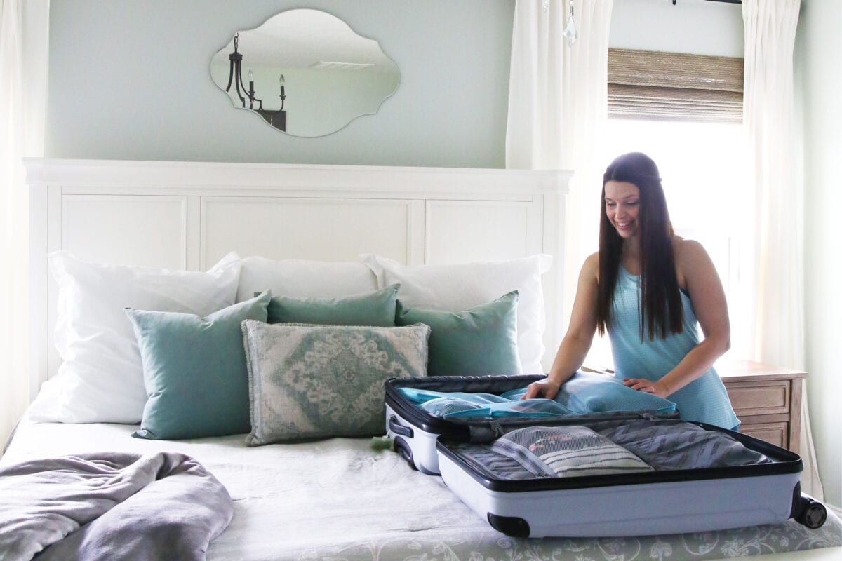 A woman in a light blue dress is smiling while packing a suitcase on a neatly made bed with blue and white pillows, ready for teaching kids about money on vacation in a bright, airy bedroom.