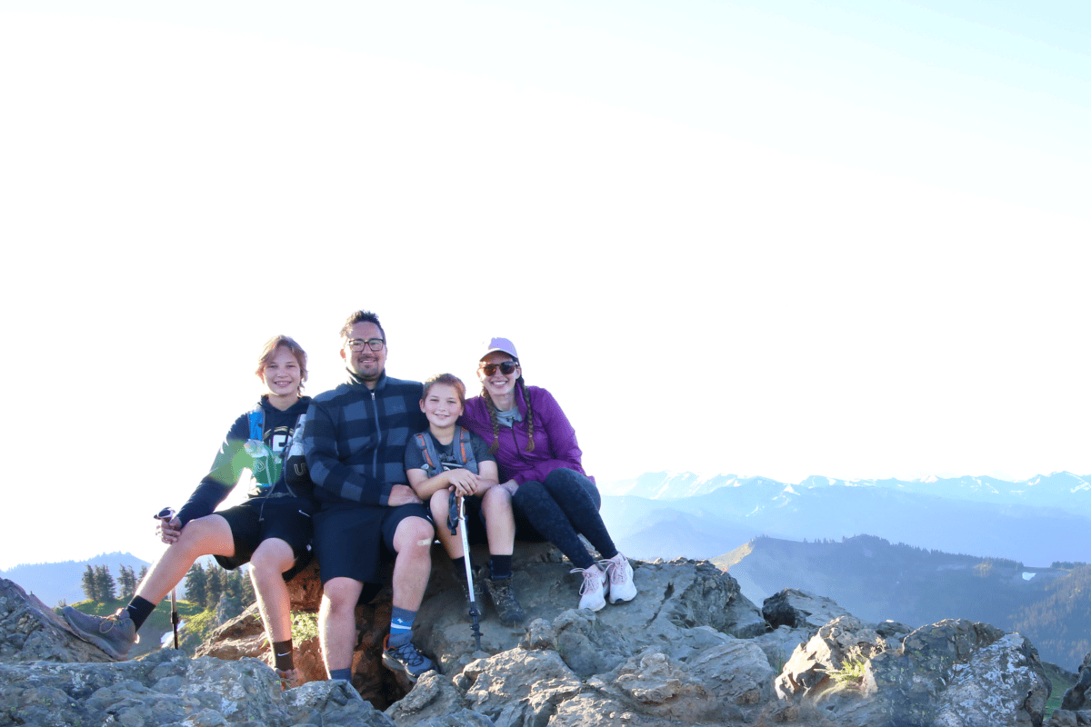 A family of four sits on rocky terrain atop a mountain, smiling at the camera. As they enjoy the view, they're also teaching kids about money on vacation, making the hike both memorable and educational.