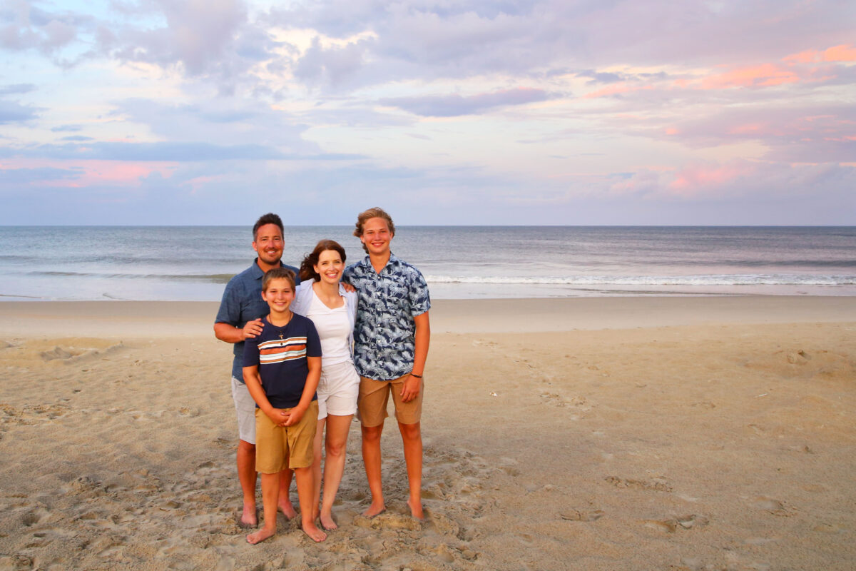 A family of four—two adults and two boys—stand smiling together on a sandy beach at sunset, enjoying the ocean and pastel sky while teaching kids about money on vacation.