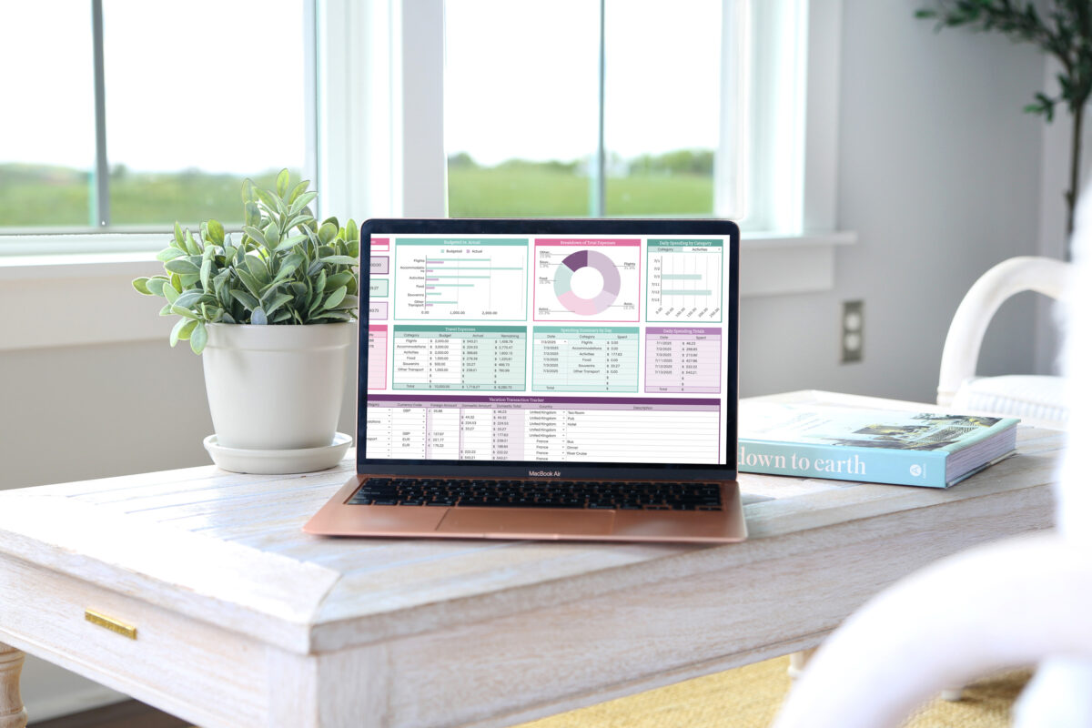 A laptop displaying financial charts and data sits on a white wooden coffee table next to a potted plant and an open book, perfect for teaching kids about money on vacation, with large windows revealing a lush green landscape in the background.