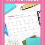A Free Printable Calendar 2027 page for January sits on a desk surrounded by pens, paper, a stapler, and teal notebooks. The calendar features a pink border with the text: "FREE PRINTABLE 2027 CALENDAR. TIME TO GET PLANNING!"—perfect for organizing your year and getting started with your plans.