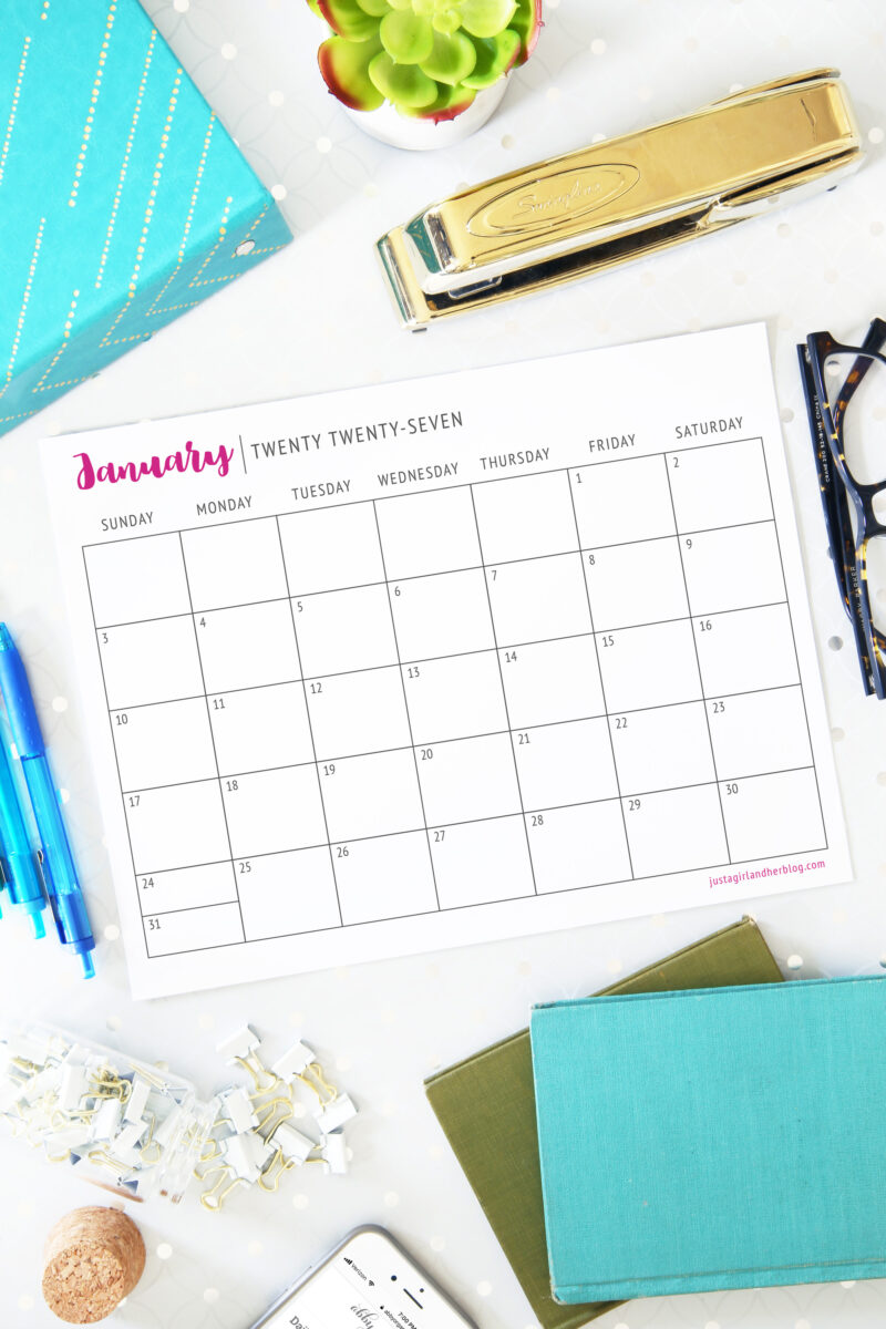 A Free Printable Calendar 2027 for January sits on a white desk, surrounded by a gold stapler, blue pen, glasses, green plant, paper clips, and teal notebooks thoughtfully arranged around it.